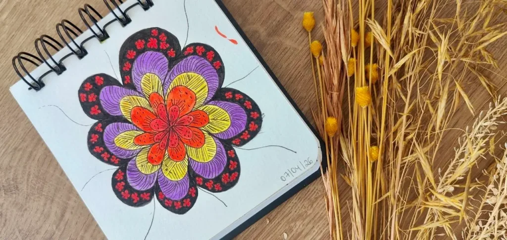 Fotografia em tons pastel e terracota, apresentando o título 'Clareza Emocional e Reorganização Energética: Atividades Manuais como Meio de Chegar ao Inconsciente'. A imagem mostra mãos segurando um pincel e colorindo uma flor estilizada (uma mandala botânica) em um caderno de anotações com espiral, representando a prática clínica e o autoconhecimento inspirado em Jung. Ao redor, há elementos simbólicos como tintas, tecidos e uma referência ao Human Design, ilustrando o processo de autodescoberta e cura.